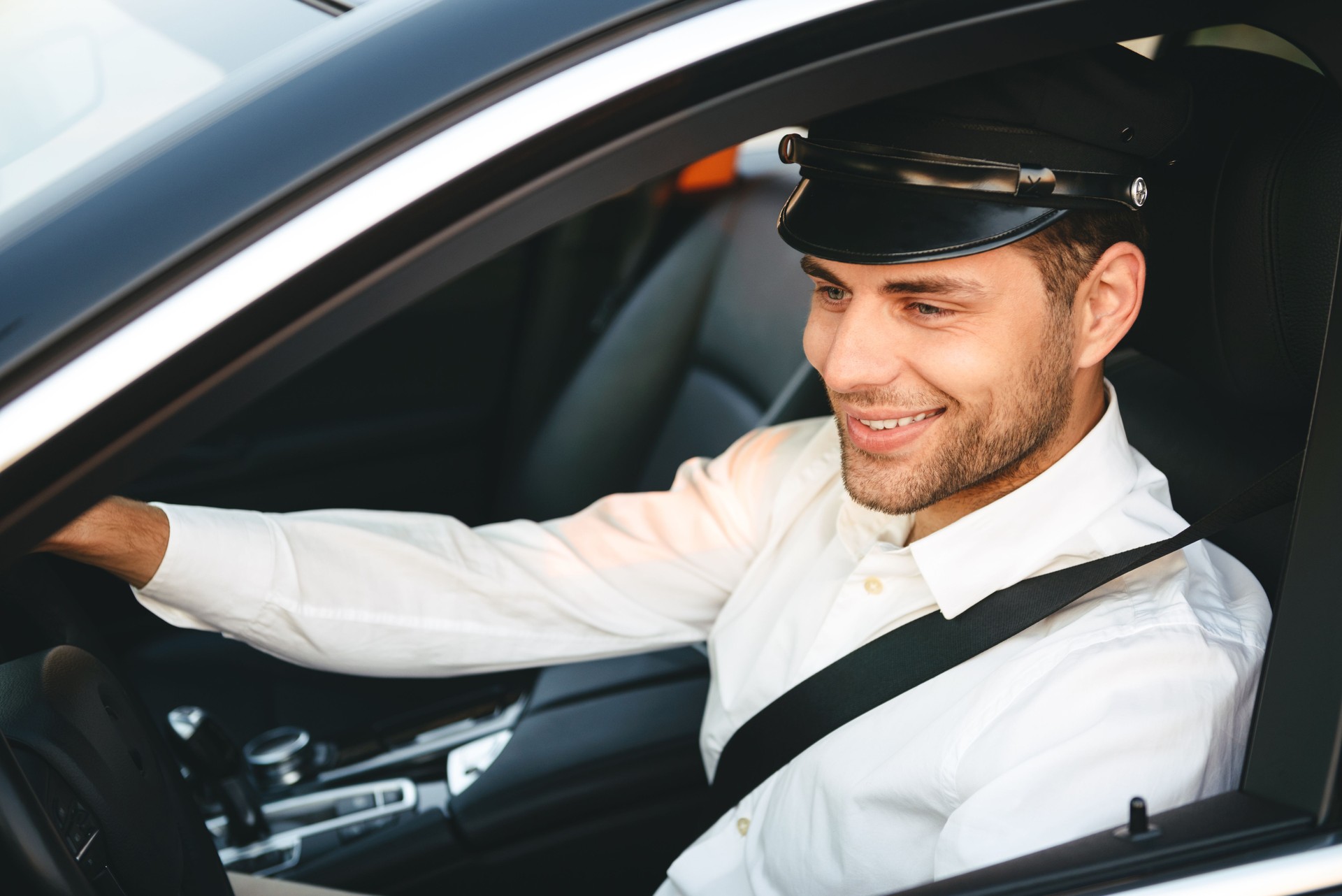 Portrait of young joyful man taxi driver in uniform and cap, driving car wearing seat belt